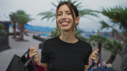 Woman holding colorful shopping bags at beach promenade under palm trees while smiling at camera; joy fun excitement spontaneity.