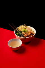 Chicken noodle bowl with vegetables, bean sprouts, and scallions served with clear soup and chopsticks on red and black background. Asian food.