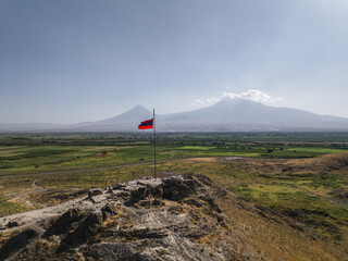 Aerial view of the Armenian flag fluttering atop a stone structure, with the distant, snow-capped Mount Ararat piercing the horizon, Khor Virap, Ararat Province, Armenia.