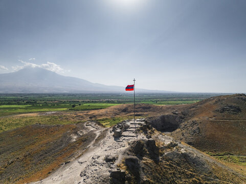 Aerial view of a red flag fluttering atop a stone structure, against a backdrop of verdant fields and distant mountains, Khor Virap, Ararat Province, Armenia.