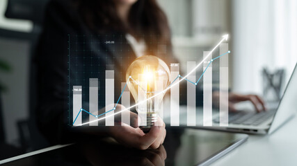 A business professional holds a light bulb symbolizing innovation while analyzing a growth chart,...
