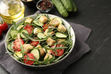Tasty salad with croutons, cucumbers, tomatoes and arugula on black table, closeup