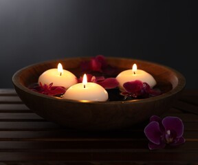 Burning candles and orchid flowers in bowl of water on wooden table against black background, closeup