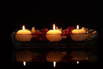 Burning candles and orchid flowers in glass container of water on mirror table against black background, closeup