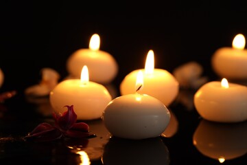 Burning candles and orchid flower on mirror table against black background, closeup