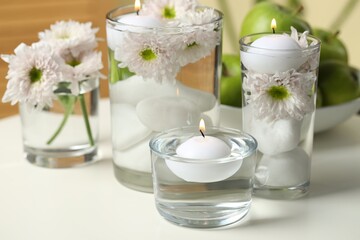 Burning candles and flowers in glasses of water on white table indoors, closeup