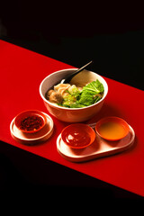 Indonesian mie ayam with chicken noodle, fresh greens, dumpling, and clear soup in ceramic bowl, Asian street food style, top view on red and black background with copy space for text.