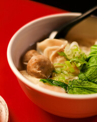 Close up of Indonesian chicken noodle soup with dumpling, meatball, and fresh greens in a white bowl. Minimalist food styling on red background.