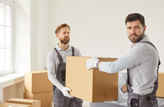 Delivery and transportation concept. Two movers carrying cardboard boxes from clients house, unload items, dressed in overalls, posing in empty room. Shipping specialists for transporting