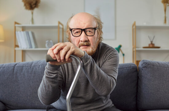 Portrait of senior man with cane at home. Old man in his 60s wearing eyeglasses sitting on sofa with hands resting on walking cane and looking at camera with unsmiling unemotional facial expression