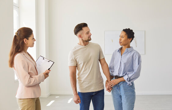 Female realtor, holding clipboard with contract, guiding young multiethnic couple through empty room during property tour. House buying, real estate, customer service, process of finding new home 