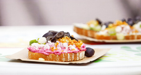 Open-faced sandwich on paper tray with creamy pink spread, sliced black olives, crumbled cheese and crispy onions, photographed on a table at an outdoor food market with soft blurred background.