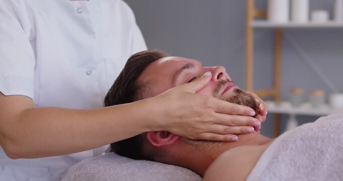 Relaxed man enjoying facial procedures at a luxury spa resort beauty salon. Side view of a handsome young man lying on a white towel while a skin care specialist does a professional face massage