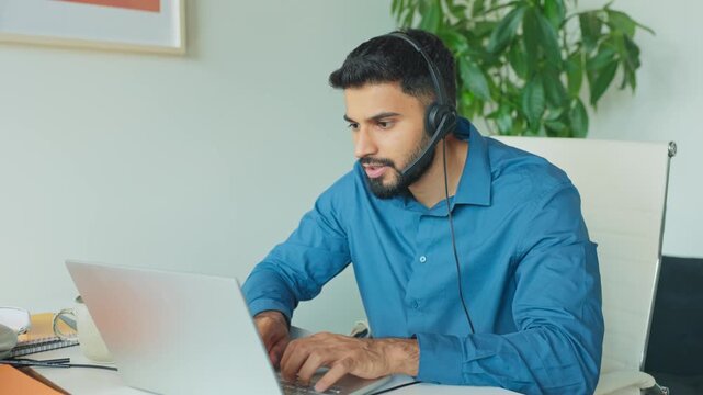 Bearded man wearing headset and working on laptop during remote call. Typing and focusing while sitting in modern office room with light walls, plants, white chair, and minimalistic workspace.