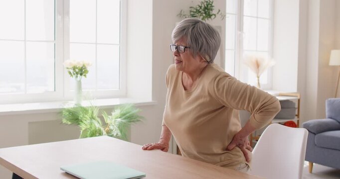 Senior woman suffers from backache and lumbar spine pain at home, feeling ache due to injury or disease while rising from table in a personal setting with focus on health challenges.