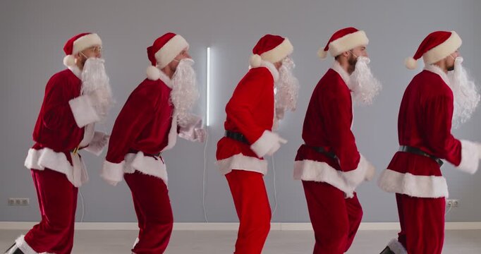Group of funny people with beard wearing Santa Claus red suits standing in a row in empty light studio stepping in place hurrying celebrate New Year and Christmas holidays. 4k video. Slow motion.