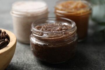 Natural body scrubs in glass jars and coffee beans on grey table, closeup
