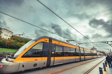 Naklejka premium A modern, streamlined tramcar in yellow and gray is moving along the tracks. Overcast skies hang above, with city buildings and people visible in the background of Porto