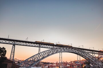 A yellow train crosses the Dom Luis I Bridge, gracefully spanning the Douro River. The sun descends, casting a warm, vibrant glow over the cityscape of Porto, Portugal