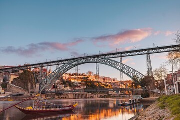 A rabelo boat gently floats on the Douro River, mirroring the golden light of the setting sun. The iconic Dom Luis I bridge stands tall, connecting the historic districts of Porto