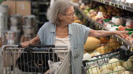 Elderly woman with gray hair carefully picks peaches in the produce section of a grocery store during the day, emphasizing health and nutrition.