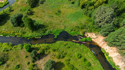 Aerial drone view of colorful kayaks floating along the winding Liswarta River, river surrounded by lush green meadows and summer nature landscape. Kayaking on river in Poland. Kayaks on a river
