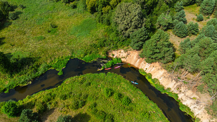 Aerial drone view of colorful kayaks floating along the winding Liswarta River, river surrounded by lush green meadows and summer nature landscape. Kayaking on river in Poland. Kayaks on a river