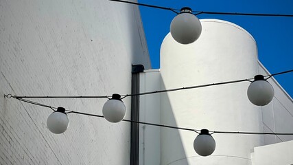 Outdoor string lighting with white globe bulbs suspended on black cables against white curved building walls and clear blue sky background