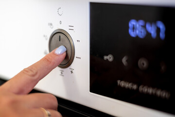 A close-up of a female hand with light blue nails adjusting a modern oven's control dial.