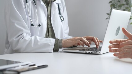 Fototapeta premium Doctor wearing lab coat and stethoscope typing on laptop keyboard and explaining something to patient during a medical consultation in hospital office. Medicine and health care concept