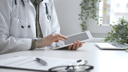 Female doctor in lab coat and stethoscope using digital tablet at desk in medical office, accessing online patient information. Medicine and health care concept