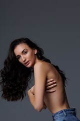 Fototapeta premium Beautiful tall thin brunette with curly hair in the studio, cyclorama. The girl stands shirtless covering her nakedness with her hands, gray background. Studio photo shoot for a model. Portrait. 