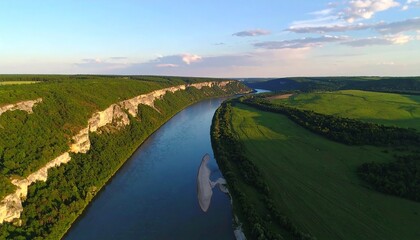 Fototapeta premium Aerial panorama of a grand river curving through a lush valley, bordered by impressive tree-covered cliffs and sprawling green fields, under a clear sky.