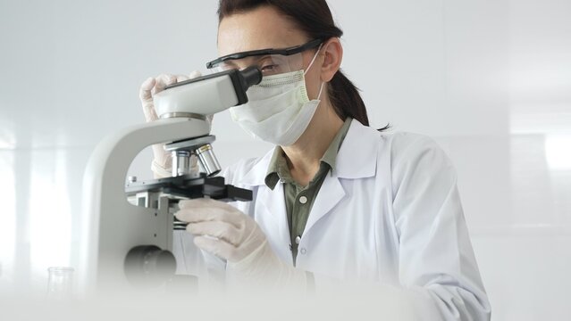 Female scientist wearing a lab coat, face mask, and safety glasses carefully adjusts a microscope, conducting research in a brightly lit laboratory setting. Medicine, healthcare and science concept