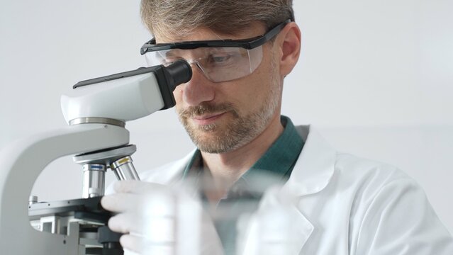 Senior man scientist wearing protective glasses and lab coat analyzing samples with a microscope in laboratory setting. Medicine, health care and science concept