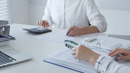 Two accountants are working together, using a magnifying glass and a calculator, to audit financial reports in their brightly lit office. Audit and taxes in business