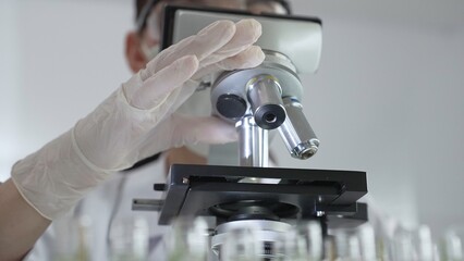 Female scientist wearing white protective gear analyzing test samples under microscope in contemporary research environment, close up of white gloved hand. Medicine and science concept