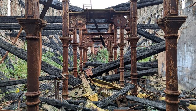 Burned down industrial factory building, charred collapsed wooden roof beams. Rusting burnt twisted metal debris. Derelict warehouse arson aftermath. Columns ruins, support pillars after fire damage.