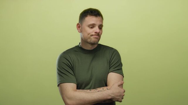 Young man with beard and tattoos stands isolated against a yellow background expressing indecision and contemplation while wearing a casual green shirt.
