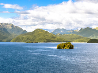 Obraz premium Serene Milford Sound with a small lush island in crystal-clear blue waters, framed by green mountains and a bright cloudy sky, New Zealand