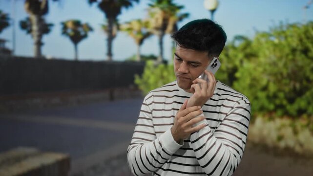Young man stands outdoors on city street using smartphone under palm trees wearing striped shirt looking thoughtful while on phone call.