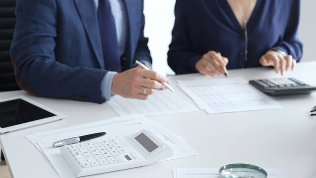 Calculator and magnifying glass over financial documents opposite financial team analyzing data, reviewing reports, collaborating at modern office workspace, close up view. Audit and taxes