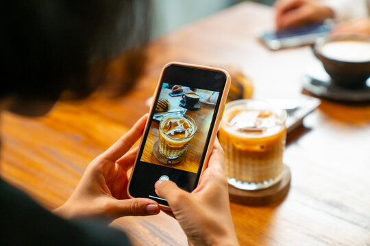Beautiful Asian woman sitting in a cozy café, enjoying coffee and pastry and using mobile phone taking picture bakery and drink on the table. Happy Woman relaxing coffee break at coffee shop.