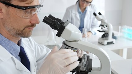 Researcher examining sample through microscope, wearing safety eyewear in sterile laboratory setting, surrounded by scientific glassware. Medicine, healthcare and science concept