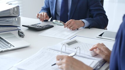Financial analysts examining documents, using magnifying glass and calculator while studying corporate data at workplace. Audit and taxes in business