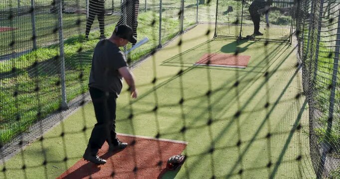 Mature adult male batter picking up bat in batting cage swinging at pitching machine for practice