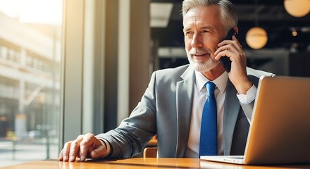 Mature businessman working in a cafe using his laptop and talking on his mobile phone near the window