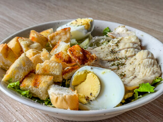 A close-up photo showing a Caesar salad dish, focusing on the sliced chicken breast, hard-boiled egg, and toasted bread (croutons) on the plate.