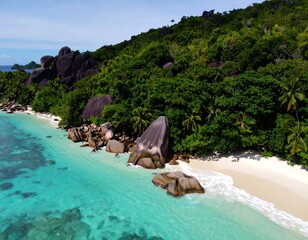 Aerial view of a tropical beach with turquoise water, white sand, and large granite boulders along a lush green hillside