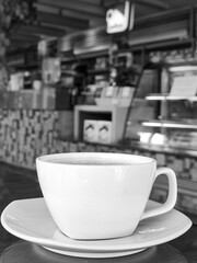 Editorial black and white photograph of a steaming coffee cup on a saucer inside a cafe environment. The blurred background with counter and menu creates a lifestyle and urban dining atmosphere.